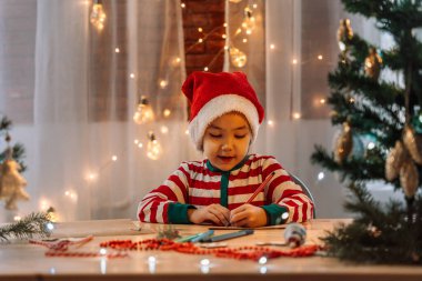 Küçük bir kız Noel Baba 'ya mektup yazıyor. Tahta bir masanın üzerine. Kalemlerin arasına ve Noel süslerinin önüne..