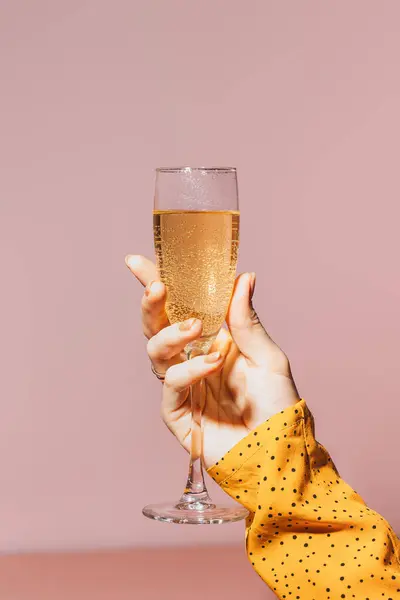 Ladys hand with gold-painted nails holding a glass of champagne against a pink background.