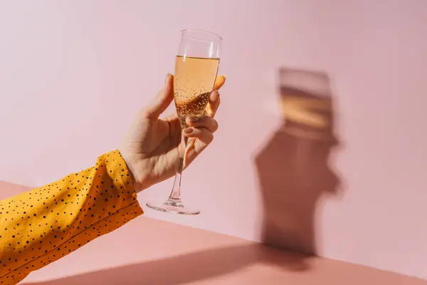 Ladys hand with gold-painted nails holding a glass of champagne against a pink background.