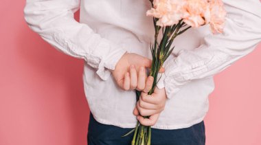 Beyaz gömlekli çocuk arkasında pembe bir arka planda bir buket çiçek tutuyor..