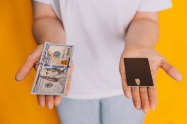 Lady holding money bills and credit card in front of yellow background. clipart