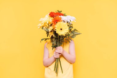 Bir çocuk sarı arka planda büyük bir buket çiçekle yüzünü kapatıyor.