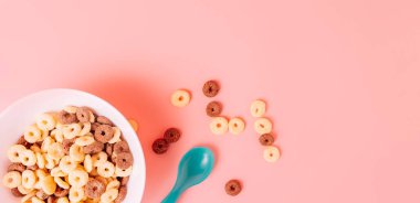 Beyaz bir tabakta sütsüz mısır halkaları. Pembe arka planda, mavi kaşıkla..