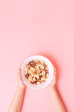 Beyaz tabakta kahvaltı gevrekleri. Pembe arka planda çocukların ellerinde..