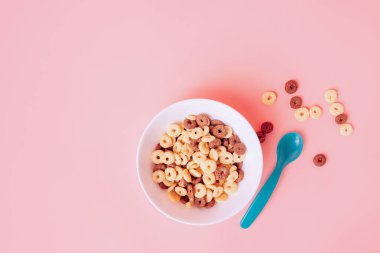 Beyaz bir tabakta sütsüz mısır halkaları. Pembe arka planda, mavi kaşıkla..