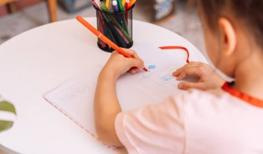 Çocuk keçeli kalemle beyaz bir masada sol elini not defterine koyuyor.
