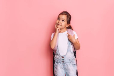 Sırt çantalı düşünceli liseli kız pembe bir arka planın önünde duruyor..