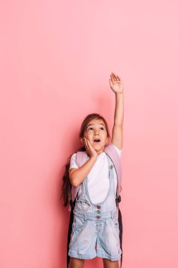 Sırt çantalı bir kız elini pembe bir arka plana kaldırdı..