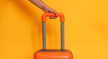 Sarı arka planda turuncu bir bavulu tutan..