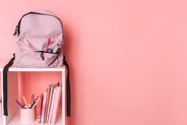 Okul malzemeleri ve beyaz raflarda pembe arka planda bir sırt çantası var..