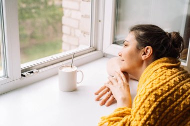 Pencerenin yanında, sarı bir kazağa sarılı bir fincan kahveyle rahatlayan bir kadın. Yüksek kalite fotoğraf