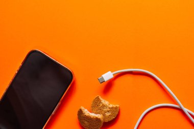 Siyah akıllı telefonun düz yatışı, beyaz kablo TV ve parlak turuncu yüzeyde yenmiş kurabiyeler dijital yaşam tarzı ve atıştırmalık pop sanat konsepti. Yüksek kalite fotoğraf
