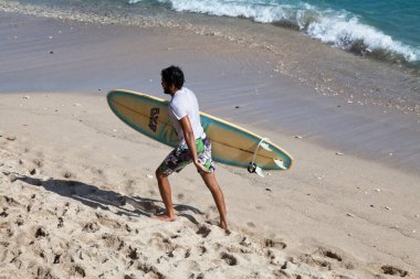 Saint-Gilles Bains, La Reunion - 13 Haziran 2017: Plage des Roches Noires sahilinde yürürken sörf tahtasını tutan genç adam).
