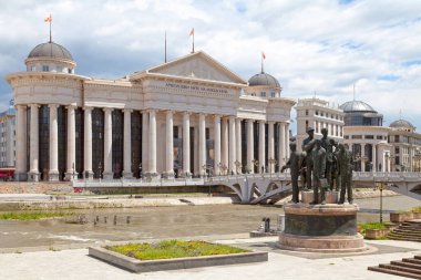 Skopje, North Mecedonia - May 21 2019: Monument of the Boatmen of Thessaloniki with the Archaeological Museum of Macedonia opposite the Bridge of Civilizations crossing the Vardar River.