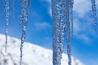 Close-up on ice stalactites formed at the bottom edge of a roof opposite a snow-capped mountain.