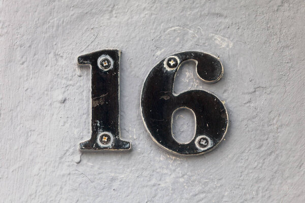Close-up on a black number 16 screwed on a concrete wall painted white.