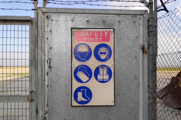 Sign attached to the metal gate of an industrial site by the sea stating: "Safety first" and showing the list of equipment required to wear to enter the premises.