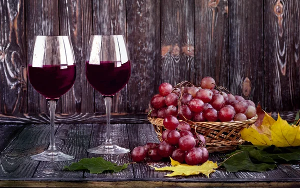 暗い木製の背景にわらバスケットに赤ワインとブドウの2つのグラス 黄色と緑の葉を持つ秋の組成 — ストック写真