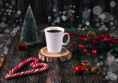 Beyaz bir fincan kahve ve koyu arkaplandaki masada kalp şeklinde kırmızı Noel lolipopları. Noel ağacı, toplar, kutsal dal, dekor olarak koniler..