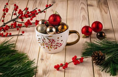 Noel topları, çam dalları, kozalaklar ve kırmızı ilex meyveleriyle dolu beyaz bir fincan. Kış, hafif ahşap arka planda sakin bir hayattır..