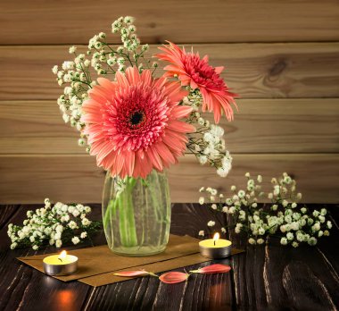 A romantic and delicate bouquet of pink gerberas in a glass vase. Gypsophila flowers, candles, flower petals, envelopes as decor on a wooden background.