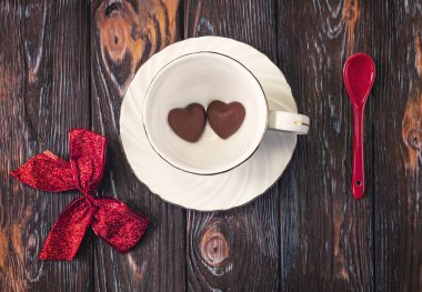 Concept for Valentine s Day. Concept for Valentine s Day. Chocolate hearts lie at the bottom of a white cup. Red bow and spoon as decor on a dark background.