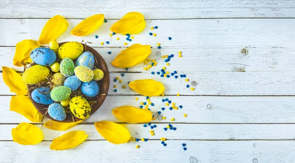 Easter background with a basket and Easter blue and yellow eggs and tulip petals on a light wooden background. Easter composition in blue and yellow colors. View from above.