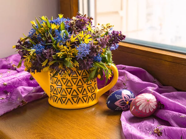 窓には青い雪の降る春の花の花束があります カラフルなイースターエッグとパープルのスカーフ 春の構図 — ストック写真