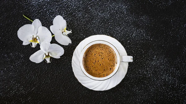White cup of coffee and white orchid flowers. Romantic composition in white on a black background. View from above