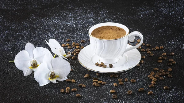 White cup of coffee, white orchid flowers, coffee beans. Romantic composition on a black background, side view.