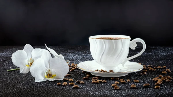 White cup of coffee, white orchid flowers, coffee beans. Romantic composition on a black background, side view.