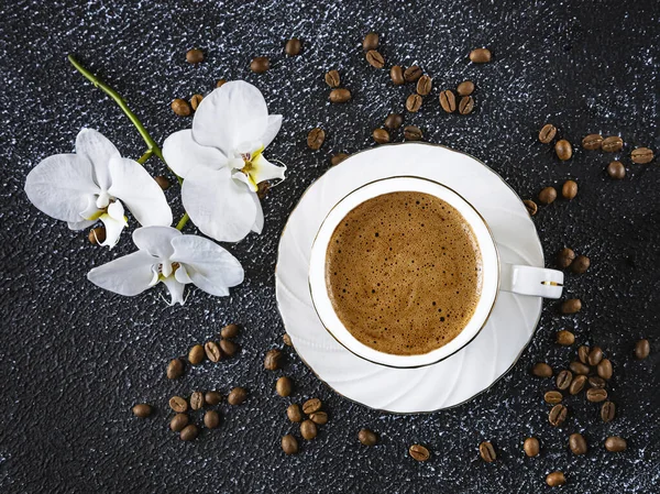 White cup of coffee, white orchid flowers, coffee beans. Romantic composition on a black background.View from above