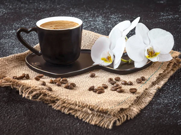 Black cup of coffee and white orchid flowers on a dark background. Coffee beans and burlap as decor. Romantic coffee composition. Side view.