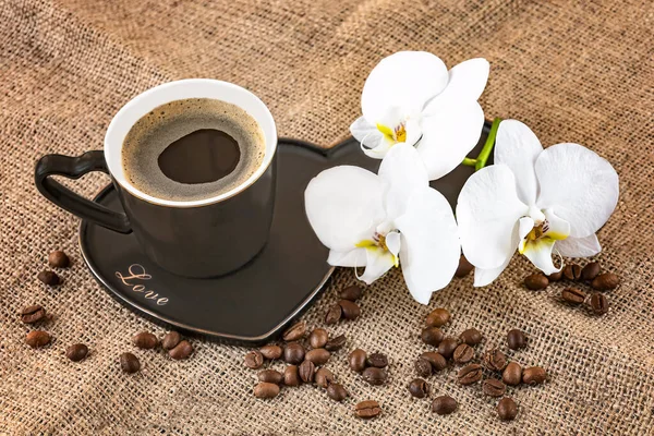 Black cup of coffee and white orchid flowers on a dark background. Coffee beans and burlap as decor. Romantic coffee composition.View from above