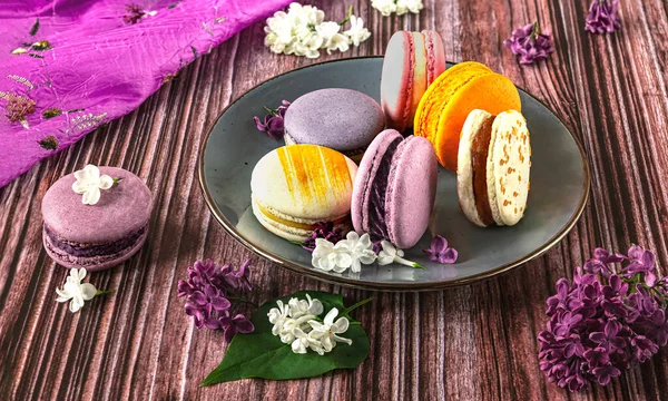 Plate with colorful French dessert macaroons on wooden background. Purple lilac flowers as decoration, side view