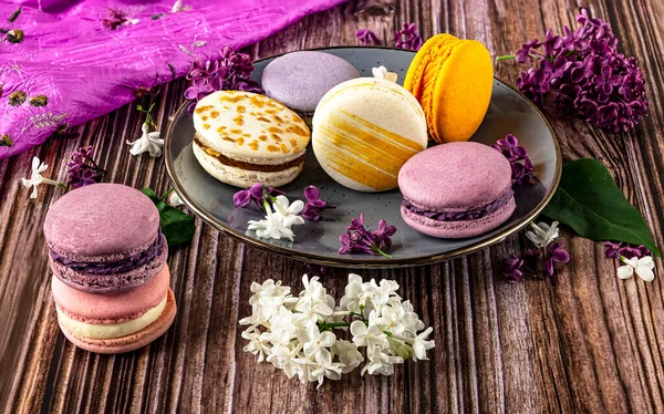 Plate with colorful French dessert macaroons on wooden background, side view. Purple lilac flowers as decoration