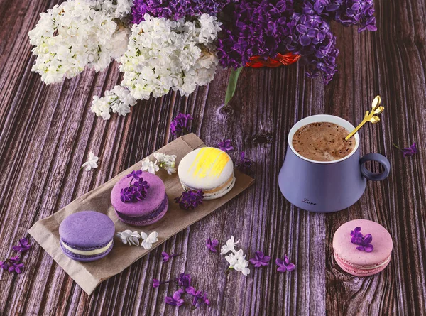 A row of colorful macaroon desserts on the table and a gray cup of coffee. Purple bouquet of lilac flowers.