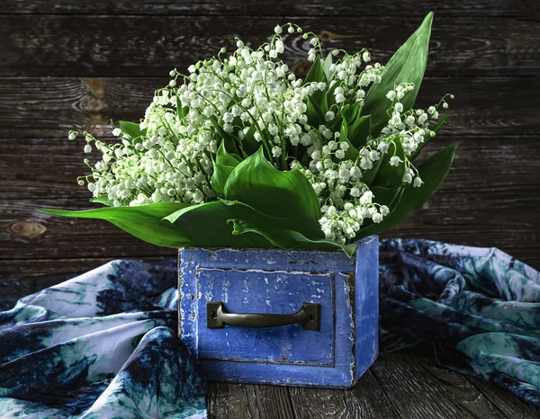 A bouquet of lily of the valley flowers in a blue wooden vintage box and a blue shawl on a wooden background. View from above.