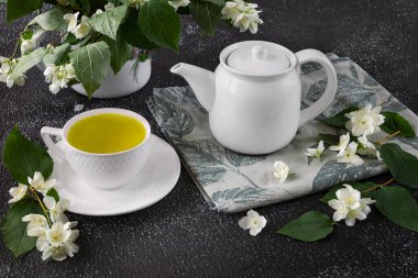 Beyaz fincanlı yeşil çay ve koyu arkaplanda çaydanlık. Süs olarak vazoda taze yasemin çiçekleri.