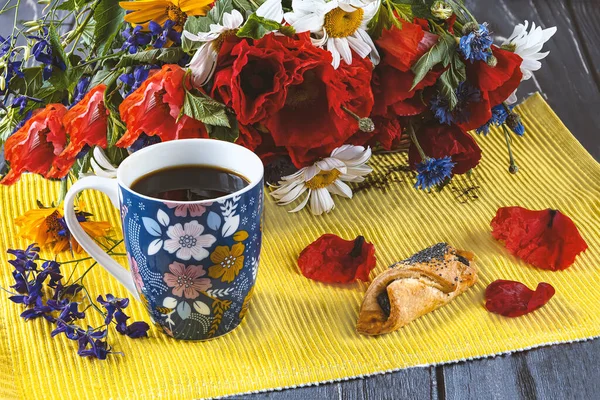 A blue cup of coffee with flowers, a bouquet of wildflowers with red poppies and cookies with poppies on a yellow background. Summer still life