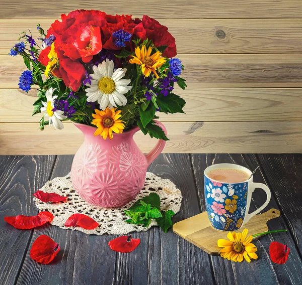 A bouquet of colorful wild flowers with red poppies in a vase, a blue cup of coffee on a wooden background. Summer still life with wild flowers