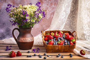 Yaz aylarında bir sepet çilek ve yaban mersini, pencere kenarında kır çiçekleri olan eski bir seramik vazo.