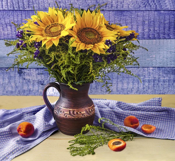 Summer still life with a bouquet of sunflowers in a clay jug, ripe apricots on a blue and yellow wooden background.