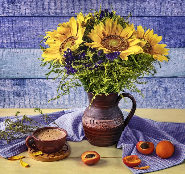 Summer still life with a bouquet of sunflowers in a clay jug, a cup of coffee, ripe apricots on a blue and yellow wooden background.