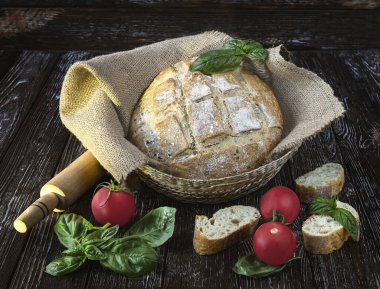 Çuval bezinde taze pişmiş çavdar ekmeği, bir parça ekmek, domates, koyu ahşap arka planda yeşil fesleğen yaprakları..