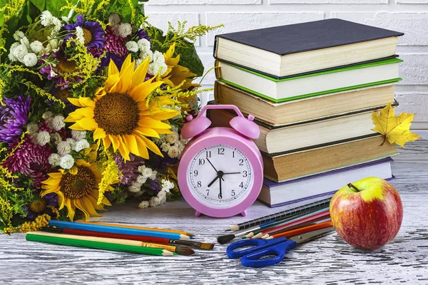 Back to school. A stack of books, a pink alarm clock, stationery, an apple, a bouquet of autumn flowers on a white background.