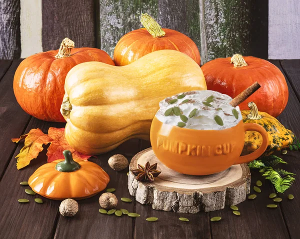 Pumpkin latte with cream, seeds, spices in a pumpkin orange cup, autumn leaves, different types of pumpkins on a wooden background. Autumn still life