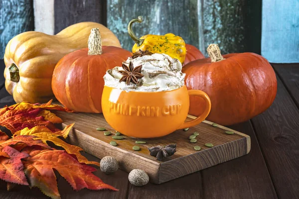 Pumpkin latte with cream and spices in a pumpkin orange cup, different types of pumpkins, autumn leaves on a wooden background.