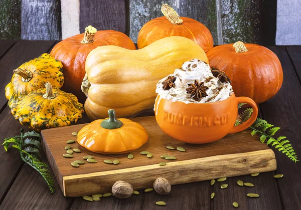 Pumpkin latte with cream and spices in a pumpkin orange cup, different types of pumpkins on a wooden background. Autumn still life