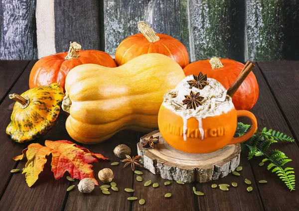 Pumpkin latte with cream and spices in a pumpkin orange cup, autumn leaves, different types of pumpkins on a wooden background. Autumn still life